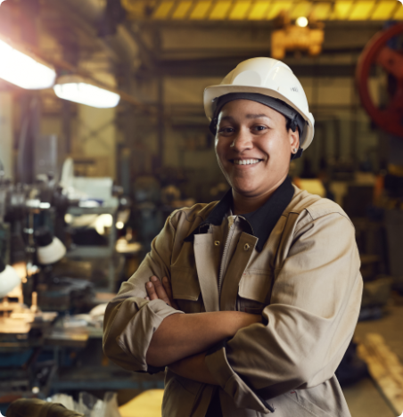 Photo : Une femme sourit fièrement. Elle porte un habit de travail, un casque de sécurité blanc et elle se trouve dans une usine.